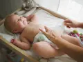 Radość noworodka podczas pielęgnacji. Mama zmienia pieluszkę, maluch uśmiecha się, leżąc na przewijaku wśród zabawek.