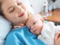 Uśmiechnięta mama tuli noworodka. To chwila spokoju po szczepieniach noworodka, budująca więź i zapewniająca zdrowie maluszka.