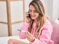 Uśmiechnięta kobieta z pozytywnym testem ciążowym rozmawia przez telefon, dzieląc się radosną nowiną.