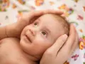 Delikatne dłonie otulają główkę dziecka, chroniąc jego miękkie ciemiączko. Maluch patrzy z zaciekawieniem w górę, uśmiechając się lekko.