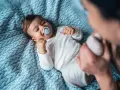 Niemowlę w białym body z niebieskim smoczkiem na niebieskim kocyku.