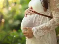 Brzuch kobiety w białej, koronkowej sukni, otoczony zielenią. Dłonie delikatnie obejmują ciążowy brzuch. W tle rozmyte światło. Luteina w ciąży wspiera rozwój maluszka.