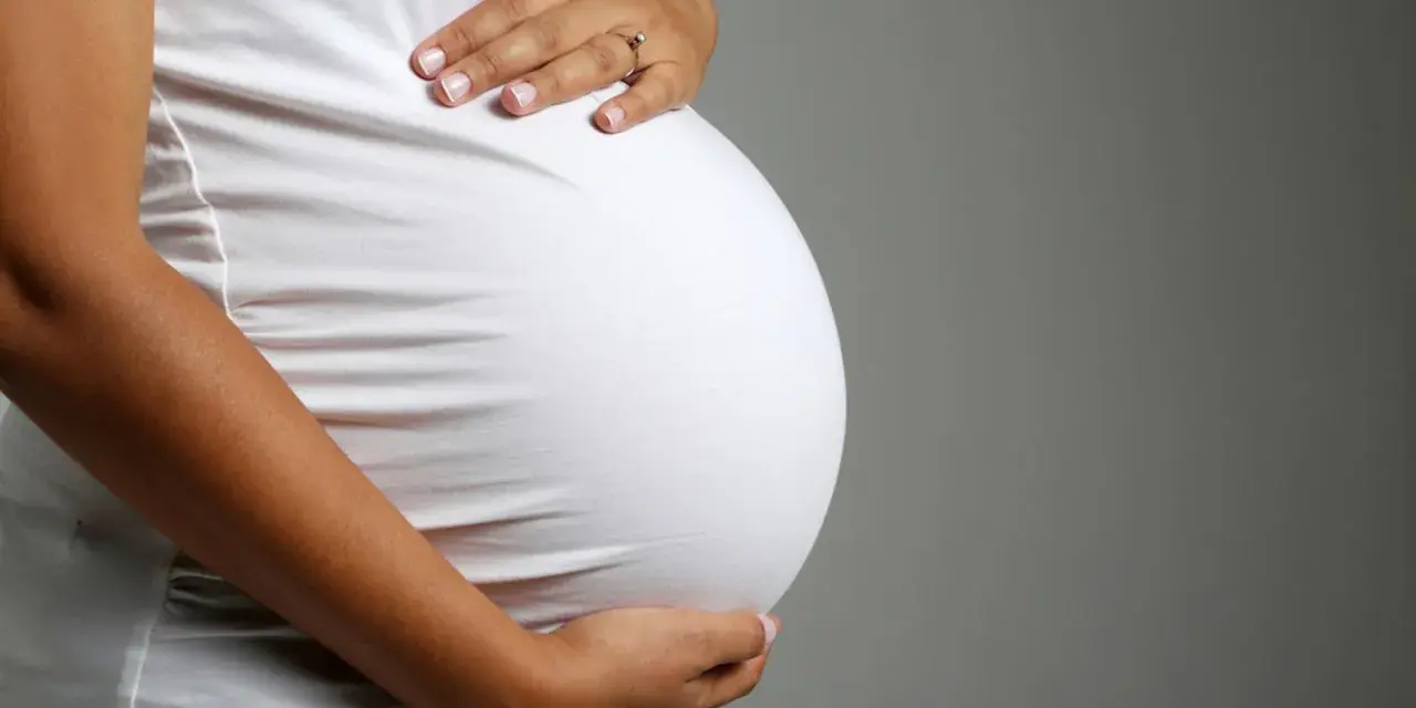 Brzuch ciężarnej kobiety w białej bluzce, dłonie delikatnie go obejmują. Zbliża się poród, może już wkrótce pojawi się czop śluzowy.