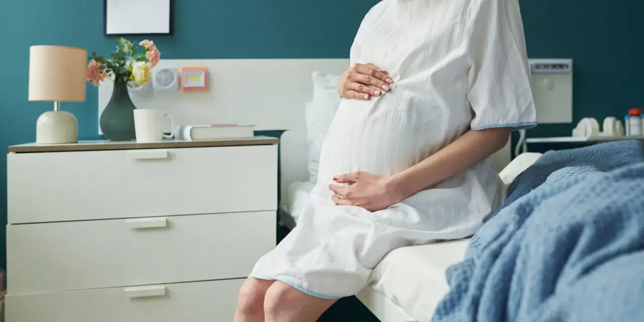 Kobieta w szpitalnej koszuli, z dłońmi na brzuchu, czeka na poród w 3 trymestrze.