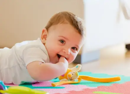 Uśmiechnięte 11 miesięczne dziecko leży na macie edukacyjnej, gryząc paluszek. Obok pluszowa zabawka.