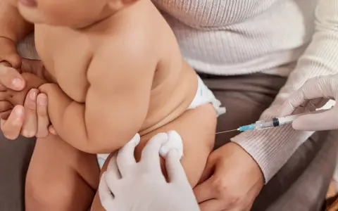 Dziecko otrzymuje szczepienie. Ręka w rękawiczce trzyma strzykawkę, a druga ręka przygotowuje miejsce wkłucia. To ważny moment dla zdrowia, jak w przypadku szczepionki heine medina.