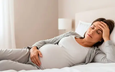 Zmęczona ciężarna odpoczywa w łóżku, czując senność w ciąży. Dłoń na czole, oczy zamknięte.