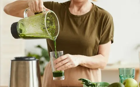 Kobieta nalewa zielony koktajl położnej do szklanki. Na blacie widać blender, awokado i jabłko.