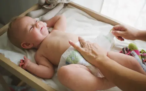 Radość noworodka podczas pielęgnacji. Mama zmienia pieluszkę, maluch uśmiecha się, leżąc na przewijaku wśród zabawek.