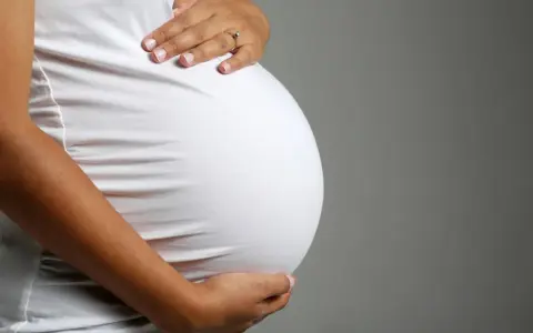 Brzuch ciężarnej kobiety w białej bluzce, dłonie delikatnie go obejmują. Zbliża się poród, może już wkrótce pojawi się czop śluzowy.