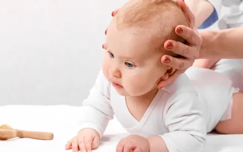 Dłoń delikatnie dotyka główki niemowlęcia, sprawdzając ciemiączko tylne. Obok leży drewniana zabawka.
