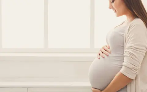 Uśmiechnięta ciężarna kobieta trzymająca się za brzuch na tle okna.