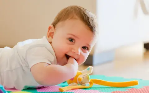 Uśmiechnięte 11 miesięczne dziecko leży na macie edukacyjnej, gryząc paluszek. Obok pluszowa zabawka.