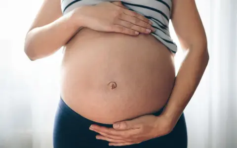 Brzuszek w ciąży z widocznym pępkiem, otoczony dłońmi przyszłej mamy.