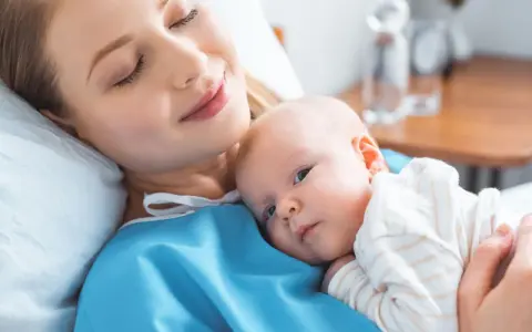 Uśmiechnięta mama tuli noworodka. To chwila spokoju po szczepieniach noworodka, budująca więź i zapewniająca zdrowie maluszka.