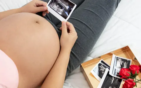 Widok z góry na ciążowy brzuch i zdjęcia USG. Widać, jak rośnie brzuch w ciąży, a obok leży pudełko z kolejnymi zdjęciami i różami.