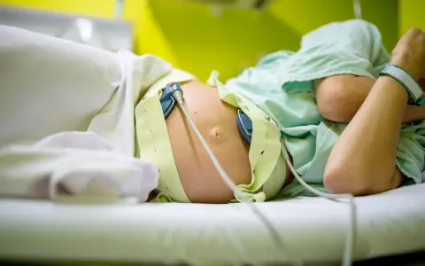 Kobieta w ciąży leży na łóżku, podłączona do aparatury KTG. Widać jej brzuch z przyczepionymi czujnikami.