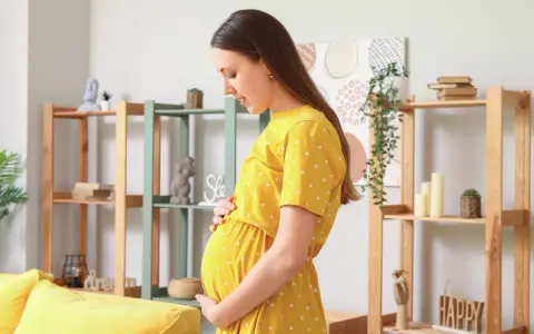 Kobieta w żółtej sukience w groszki czule trzyma brzuch. W tle regały z dekoracjami, sugerujące domowe ciepło i oczekiwanie na dziecko, być może zmagająca się z wielowodziem.