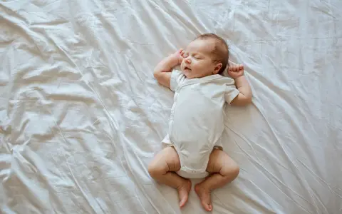 Śpiący, 3 tygodniowy noworodek w białym body leży na pogniecionej białej pościeli.