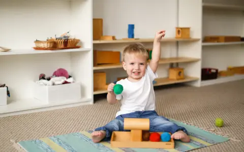 Uśmiechnięte dziecko 2 lata z zieloną kulką w dłoni, siedzi na dywanie obok drewnianej zabawki z kolorowymi kulkami.