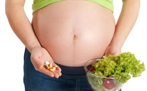 Kobieta w ciąży trzyma garść tabletek i miskę sałatki. Ważne jest żelazo dla kobiet w ciąży, czy wybrać suplementy czy zdrową dietę?