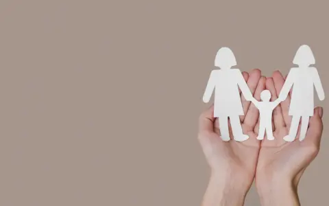 Dwie dłonie trzymają białe wycinanki: dwie postacie dorosłych i między nimi tęczowe dziecko.