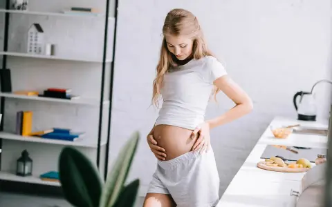 Kobieta w ciąży, odczuwająca duszności, delikatnie dotyka brzucha. W tle kuchnia i regał z książkami.