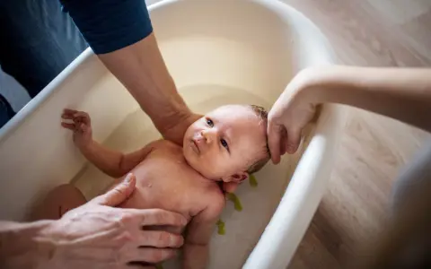 Delikatna kąpiel noworodka w wanience. Rodzice ostrożnie trzymają dziecko, dbając o jego komfort. Woda jest ciepła, a kąpiel przypomina kojący **krochmal do kąpieli**.