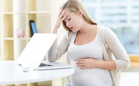 Zmęczona ciężarna z dłońmi na czole i brzuchu, czuje kołatanie serca w ciąży przy laptopie.
