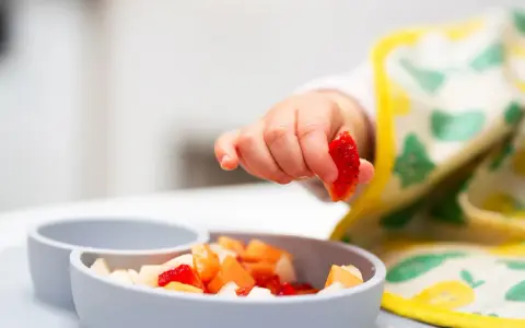 Rączka 9 miesięcznego dziecka sięga po kawałek truskawki z miseczki pełnej owoców.