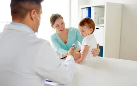 Pediatra bada niemowlę podczas pierwszej wizyty. Mama z uśmiechem obserwuje badanie.