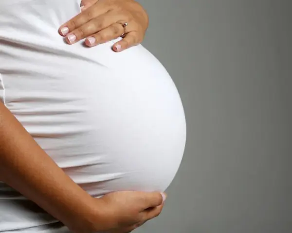 Brzuch ciężarnej kobiety w białej bluzce, dłonie delikatnie go obejmują. Zbliża się poród, może już wkrótce pojawi się czop śluzowy.