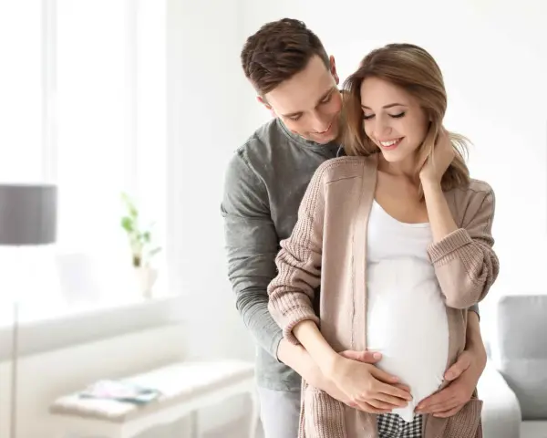 Szczęśliwy mąż obejmuje żonę w ciąży, okazując troskę i miłość. To piękny obraz wsparcia i oczekiwania na dziecko, symbolizujący opiekę na żonę po porodzie.