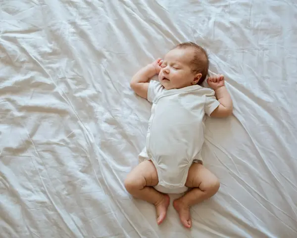 Śpiący, 3 tygodniowy noworodek w białym body leży na pogniecionej białej pościeli.
