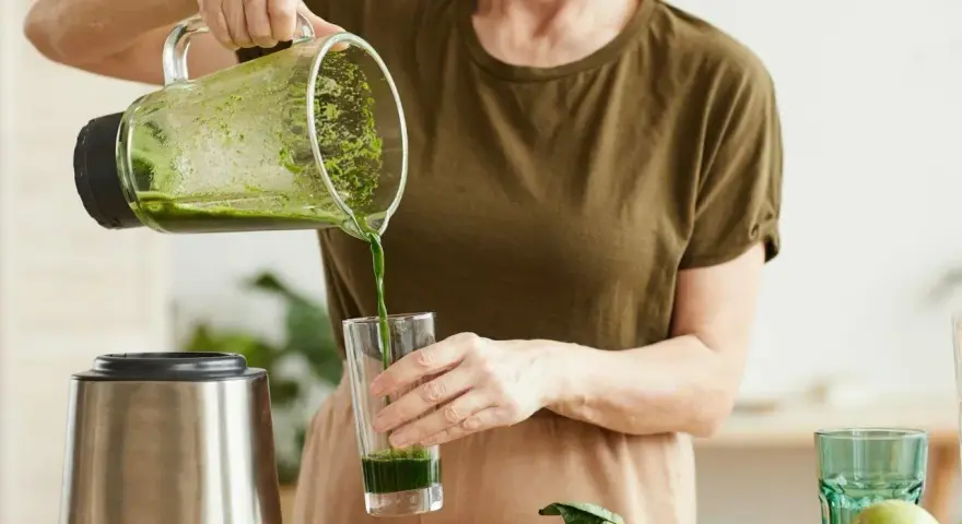 Kobieta nalewa zielony koktajl położnej do szklanki. Na blacie widać blender, awokado i jabłko.