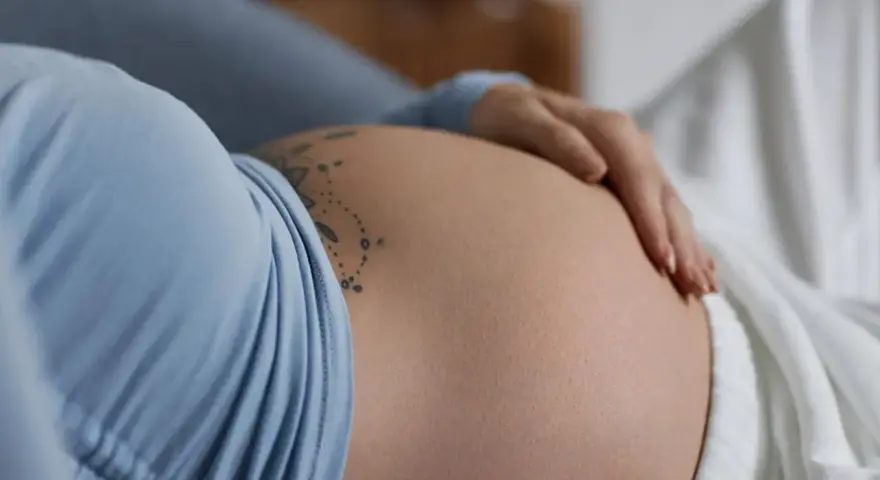 Dłoń delikatnie głaszcze ciążowy, twardy brzuch z tatuażem.