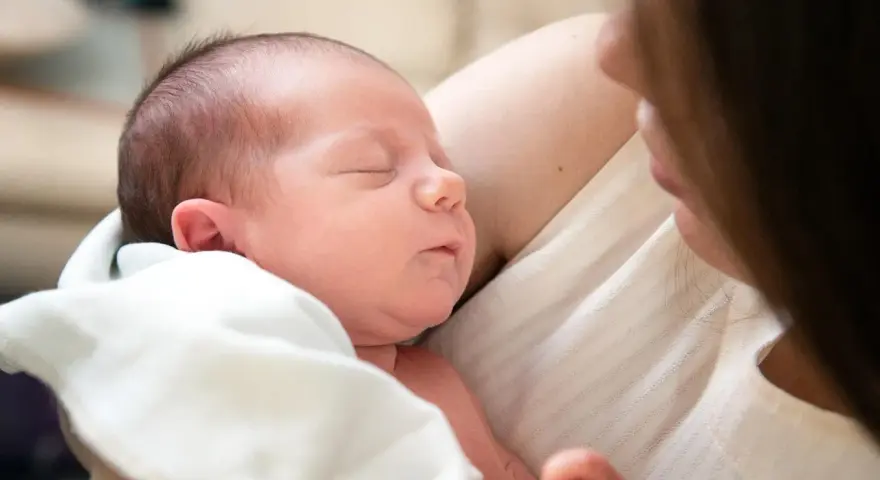 Śpiący noworodek wtulony w ramiona mamy. Delikatne światło podkreśla jego rysy.