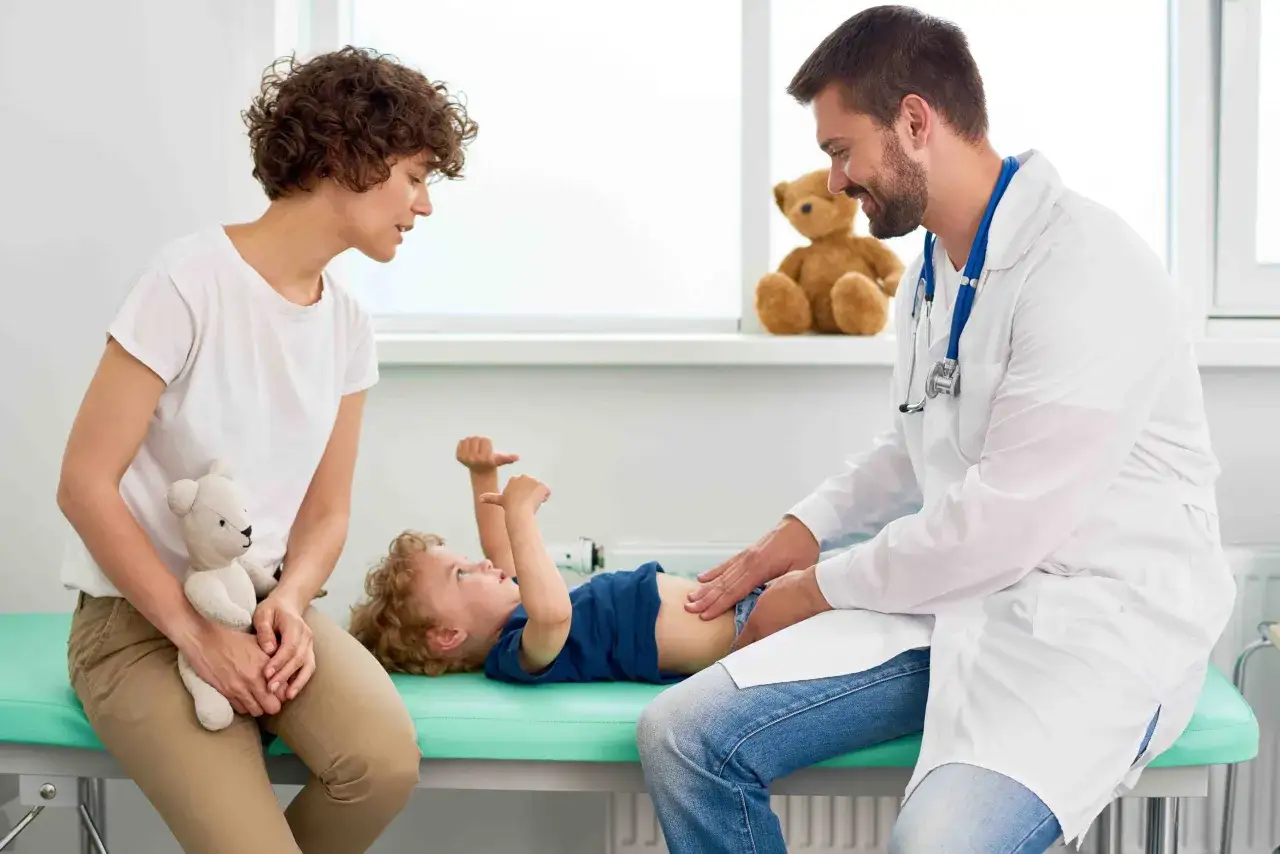 Lekarz bada brzuszek dziecka, które ma problemy z wymiotami. Obok siedzi zaniepokojona mama z misiem.