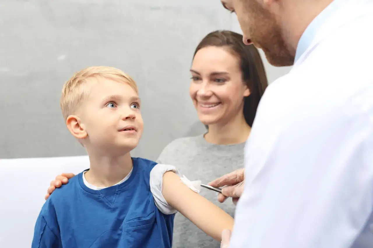 Uśmiechnięta mama wspiera swojego 5-latka podczas szczepienia. Lekarz w rękawiczkach przygotowuje ramię chłopca.