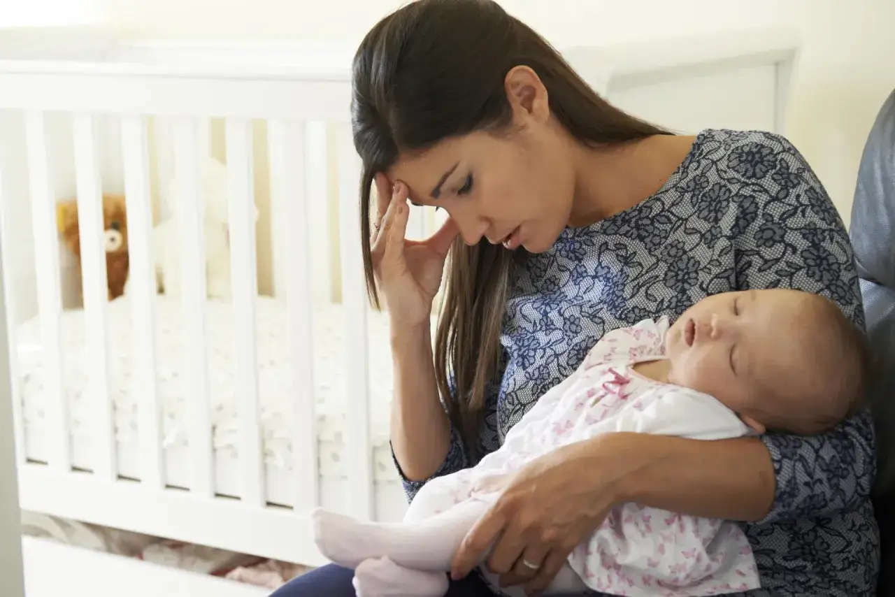 Zmęczona mama tuli śpiące dziecko, czując przygnębienie i baby blues. W tle białe łóżeczko.